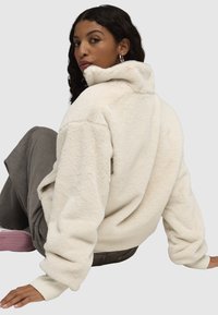 Cream-colored fleece jacket with a high collar, soft texture, and ribbed knit cuffs. Model seated, wearing olive pants and pink shoes.