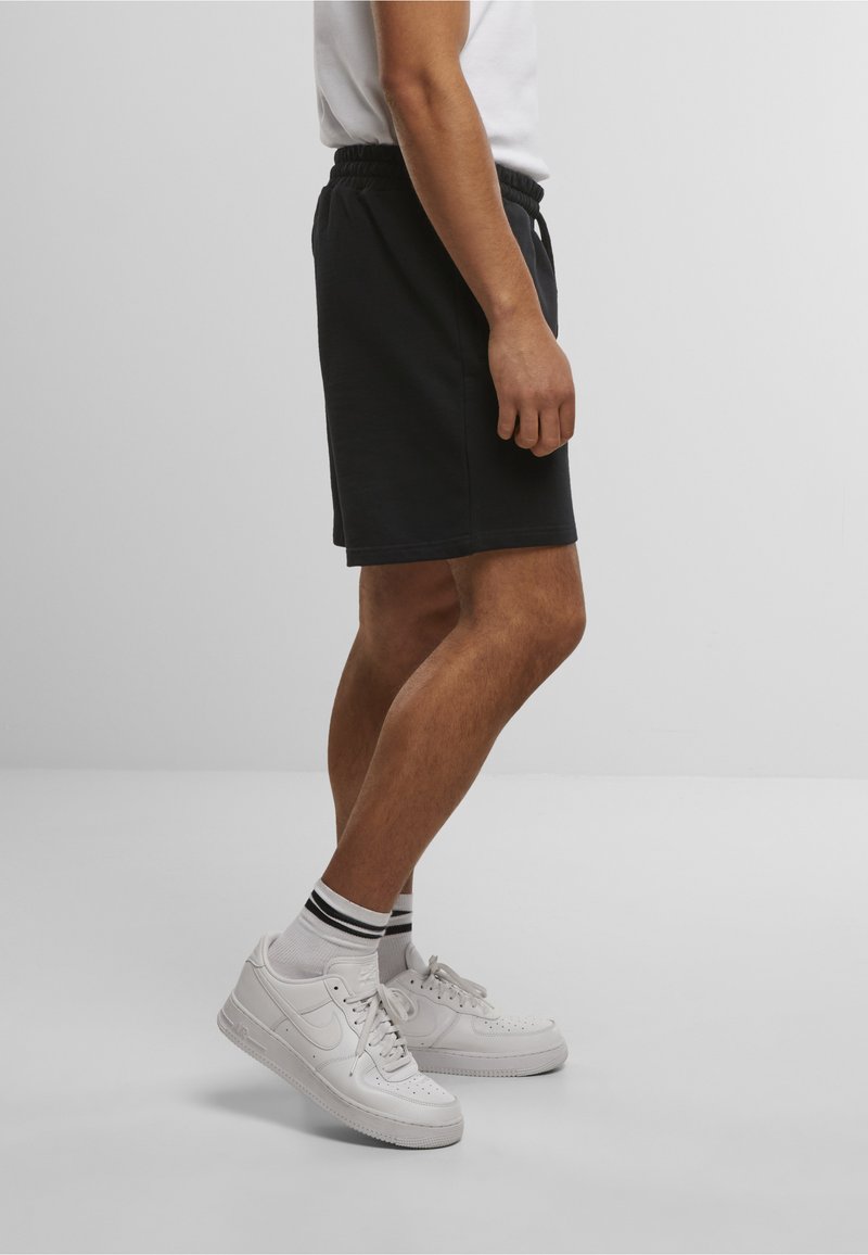 Mand iført sorte shorts med elastisk talje, hvide sneakers og hvide sokker med sorte striber på en ensfarvet baggrund.
