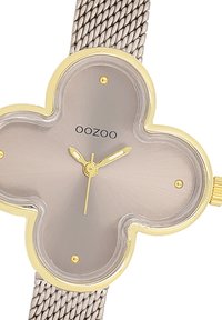 Montre à quartz avec un boîtier en forme de trèfle doré, un cadran gris et un bracelet en maille. Inclut des accents dorés subtils et des repères d'heure clairs.