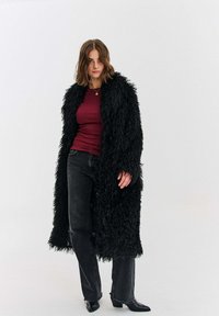 Černý, nadýchaný dlouhý kabát s texturovaným povrchem. Nošený přes vínově červenou košili, doplněný černými džíny a botami s mírným podpatkem.