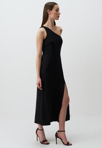 Abito nero senza maniche con un design asimmetrico e uno spacco alto fino alla coscia. Il materiale sembra liscio, abbinato a sandali neri con cinturini e tacco alto.