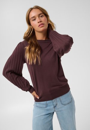 Woman with long blonde hair wearing a dark purple ribbed sweater with puffed sleeves and light blue jeans against a plain background.
