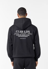 Άνδρας που φοράει μαύρο φούτερ με κουκούλα με λευκό κείμενο "CLSR LDN Luxury British Atelier Closure Design Studio Crafted in Soho Established 2015" στην πλάτη.