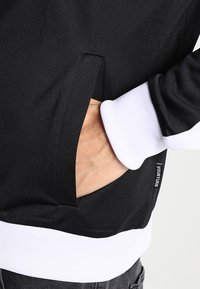 Black sweatshirt featuring a white ribbed cuff and waistband, with a hidden side pocket and a tag reading "YOUR TURN" on the hem.