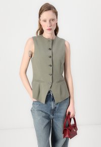 Oliven grønn ermeløs vest med fem knapper, to frontlommer og en skreddersydd silhuett, parret med blå denimjeans og en liten rød håndveske.