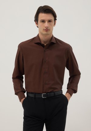 Man met donker haar die een bruin overhemd met knopen draagt, ingestopt in een zwarte broek met een zwarte riem, staande met zijn handen in zijn zakken.