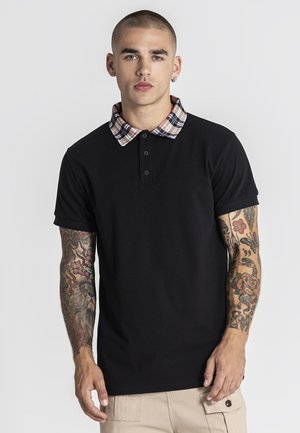 Giovane uomo con tatuaggi su entrambe le braccia che indossa una maglietta polo nera con colletto a quadri e pantaloni beige su uno sfondo semplice.