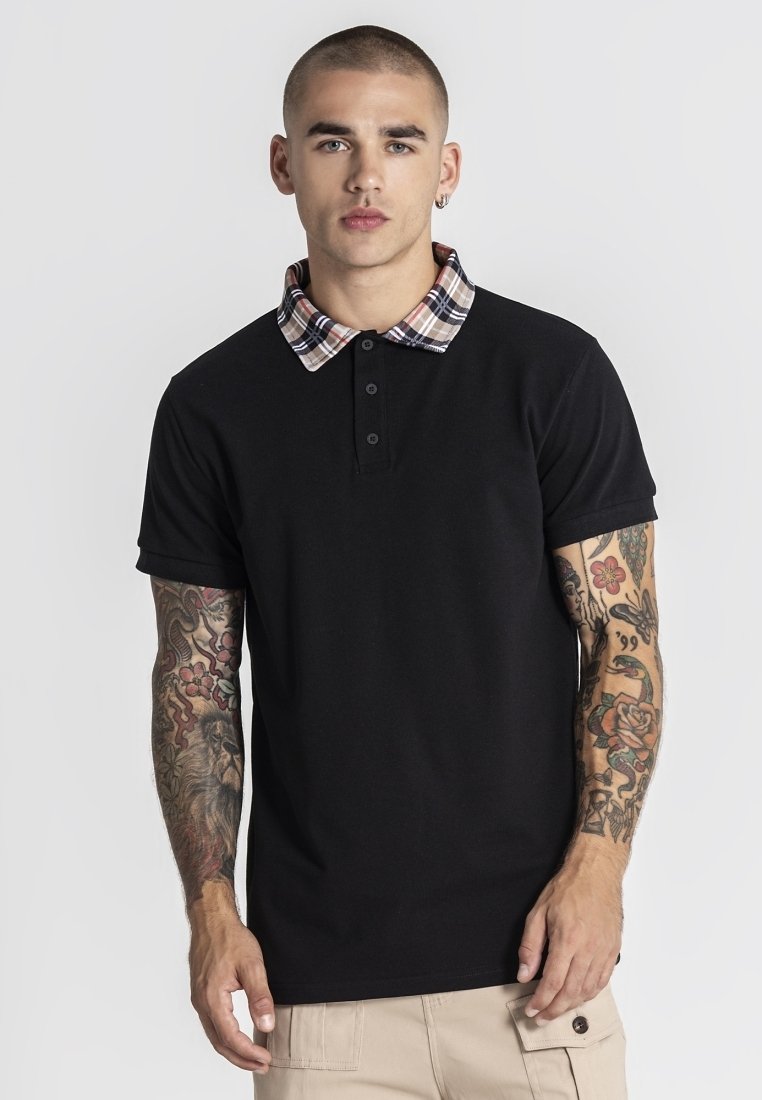 Junger Mann mit Tätowierungen auf beiden Armen, trägt ein schwarzes Polohemd mit karierter Kragen und beige Hose vor einem einfarbigen Hintergrund.