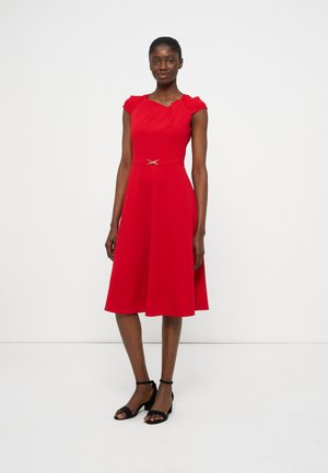 Γυναίκα στέκεται φορώντας ένα κόκκινο φόρεμα μήκους μέχρι το γόνατο με κοντά μανίκια και μια μικρή ζώνη στη μέση, συνδυασμένο με μαύρα σανδάλια με λουράκι στον αστράγαλο.