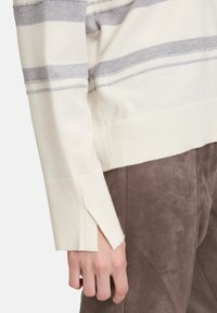 Cremfarbener Pullover mit grauen Streifen, gerippten Bündchen und seitlichen Schlitzen. Weicher Stoff, körperbetonter Schnitt. Trägt man über braunen Wildlederhosen.