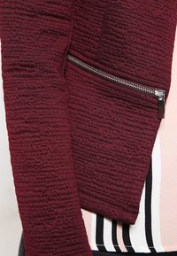 Veste bordeaux en tissu texturé, avec une poche latérale zippée et des lignes épurées. En arrière-plan, un haut à motifs rayés.