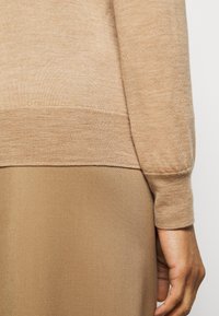 Beige Pullover aus weichem Material, mit einem taillierten Bündchen und Saum. Kombiniert mit einer glatten, leicht glänzenden beigen Unterbekleidung.