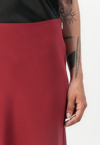 Jupe bordeaux en tissu lisse avec une fermeture éclair sur le côté. Présente un design épuré et une silhouette ajustée, reposant juste au-dessus de la taille.