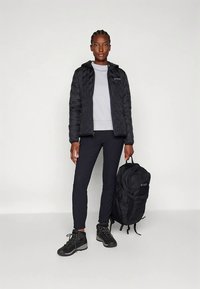 Žena u crnoj pritiskanjoj jakni, crnim hlačama, planinskim čizmama, drži crni ruksak uzbijeli pozadinu.