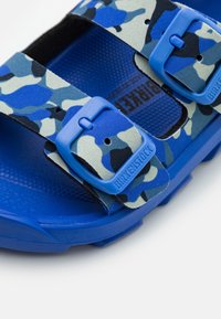 Sininen camo-kuosisandaali, jossa on kaksi säädettävää hihnaa, kumipohja ja Birkenstock-logot. Teksturoiduilla pohjilla tuki.