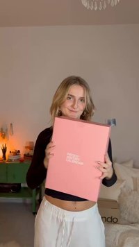 Roze rechthoekige doos met witte tekst, vastgehouden door een persoon. De doos heeft een gladde afwerking, met een minimalistisch ontwerp en geen zichtbare patronen.