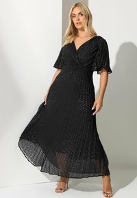 Kobieta ubrana w czarną plisowaną sukienkę midi z falbaniastymi rękawami i subtelnymi kropeczkami, w połączeniu ze złotymi sandałami na obcasie, pozująca z wdziękiem.