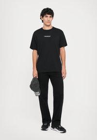 Svart oversized t-skjorte med hvit logo på brystet, kombinert med svarte bukser og svarte sneakers. Holder en grå cap med hvit tekst.