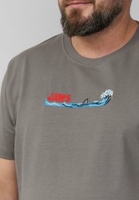 Grijze katoen T-shirt met rode 'JAWS' tekst, blauwe geborduurde golven en een haaienvin ontwerp, met een ronde hals en korte mouwen.