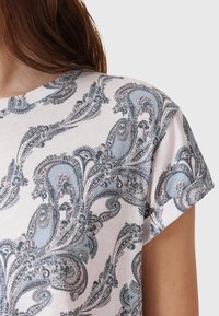 Krótki rękaw T-shirt z miękkiego materiału z kwiatowym wzorem paisley w jasnoniebieskim, różowym i ciemnoniebieskim na kremowym tle.