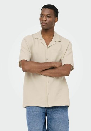 Mand med korslagte arme iført kortærmet beige skjorte med knapper og blå jeans, stående mod en ensartet lys baggrund.