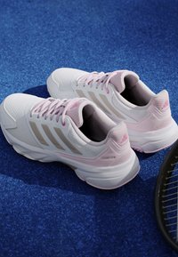 Hvide og lyserøde Adidas tennissko med snørebånd på en blåt struktureret overflade, ved siden af en sort tennisketsjerramme.