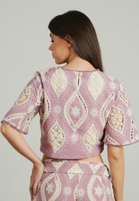 Rosa Cropped-Top mit Häkelspitze in Blumen- und Rautenmuster. Kurze Ärmel und ein Knopfverschluss auf der Rückseite.
