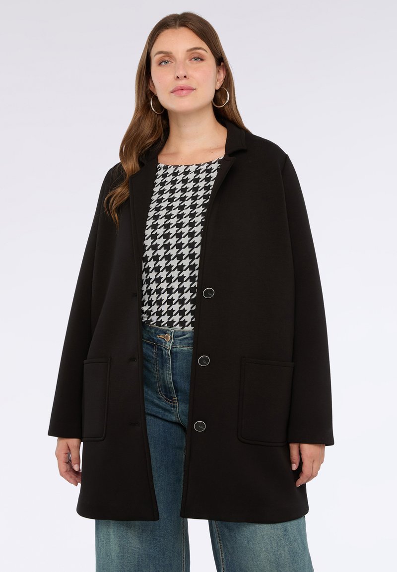 Abrigo negro, de gran tamaño, con un diseño con cuello, dos bolsillos frontales y herrajes plateados. Lleva puesto sobre una blusa de patrón de espiga blanco y negro.