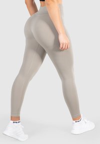 Hochgeschnittene, beige Leggings aus einem glatten, dehnbaren Stoff mit seitlichen Nähten und Knöchelbündchen. Getragen mit weißen Sportschuhen.