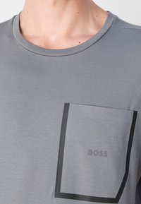 Gros plan d’un t-shirt gris avec un col rond et une poche poitrine rectangulaire bordée de noir, avec le mot BOSS.