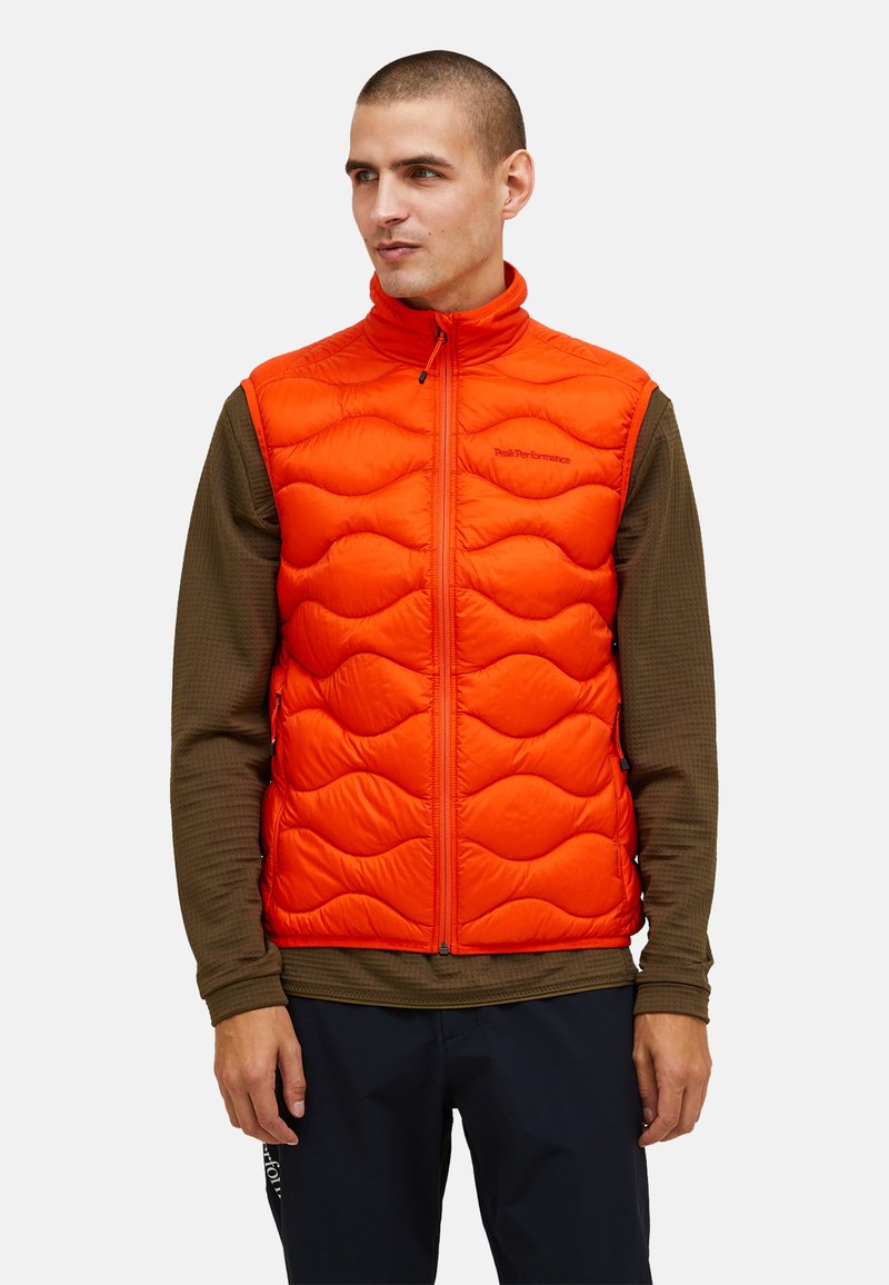 Gilet imbottito arancione brillante con colletto alto, cerniera completa e tasche laterali. Indossato sopra una camicia a maniche lunghe marrone texture.