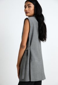 Nainen, jolla on pitkät mustat hiukset ja yllään hihaton harmaa raitapaita, seisoo sivuttain ja katsoo yli olkansa yksinkertaista taustaa vasten.