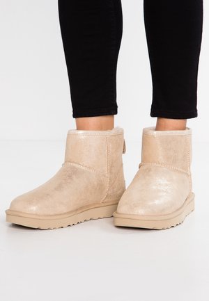 Paar beige winterlaarzen tot halverwege de kuit met een zachte voering, gedragen met zwarte nauwsluitende broek, tegen een lichte vloer- en muurachtergrond.