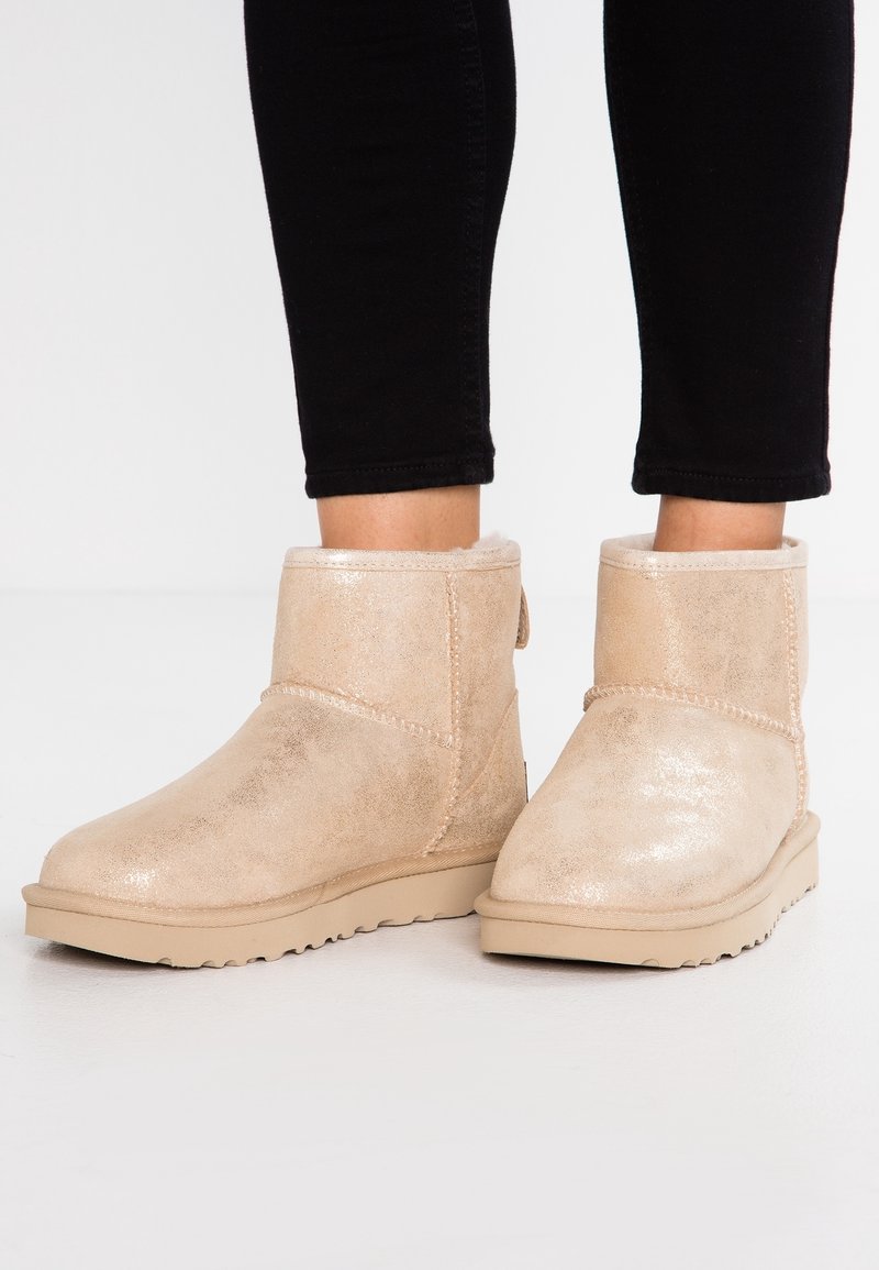 Paire de bottes d'hiver beiges mi-mollet avec une doublure moelleuse portées avec un pantalon noir ajusté sur un sol et un mur clairs.