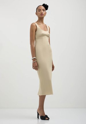 Abito beige aderente fino al ginocchio con scollatura a barca e spalline larghe. Tessuto liscio. Indossato con sandali neri con tacco alto e un accessorio per capelli floreale.