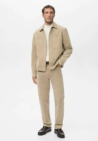 Chaqueta de lona beige, cuello alto blanco, pantalones beige a juego y zapatos oscuros. La chaqueta tiene dos bolsillos frontales. Diseño sencillo y ajustado.