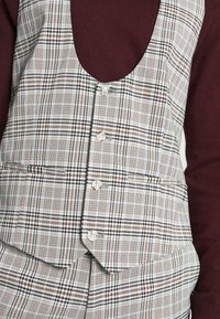 Ruudussa beige- ja ruskeasävyt. Sisältää syvän kaula-aukon, muotoiltua istuvuutta, viisi nappia ja etutaskun käytännöllisyyttä varten.