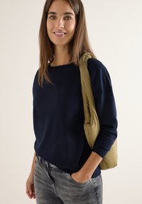 Maglione lavorato a maglia blu navy, vestibilità rilassata, scollatura a girocollo. Jeans grigi con tasche. Borsa trapuntata verde oliva appoggiata sulla spalla.
