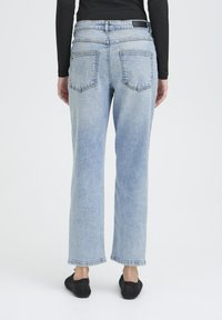 Jeans de mezclilla azul claro con un diseño de pierna recta, que presenta dos bolsillos traseros y un dobladillo deshilachado. Combinados con zapatos negros de tipo mocasín.