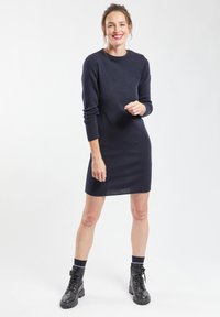 Marineblauwe gebreide jurk met lange mouwen, een ronde hals en knoopdetails op de schouders. Draagt bij zwarte veterschoenen en marineblauwe sokken.