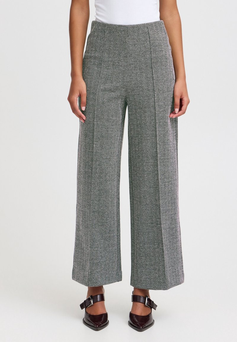 Femme portant un pantalon gris en chevrons à taille haute et jambes larges, et des chaussures noires pointues avec des brides à la cheville, debout devant un fond blanc.