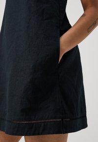 Marineblaues Leinenkleid mit entspannter Passform, seitlichen Taschen und einem Saum mit subtilen Ausschnitten. Glatte Textur und leichtes Material.