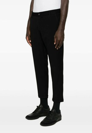 Man die zwarte cropped broeken, zwarte sokken en zwarte sneakers draagt, staande tegen een effen witte achtergrond.