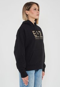 Felpa nera con cappuccio e logo dorato, tasca a marsupio, polsini e orlo a costine; texture liscia, vestibilità rilassata. Indossata con jeans azzurri.