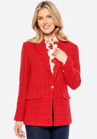 Blazer in tweed rosso con motivo a quadri, chiusura con un singolo bottone dorato e tasche con pattina, indossato sopra una blusa floreale, abbinato a jeans scuri.