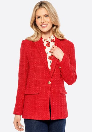 Blazer in tweed rosso con motivo a quadri, chiusura con un singolo bottone dorato e tasche con pattina, indossato sopra una blusa floreale, abbinato a jeans scuri.