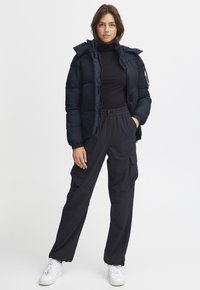 Marineblauwe pufferjack met capuchon, ritsluiting en gestructureerde stof; gecombineerd met losse zwarte cargobroek en witte sneakers.