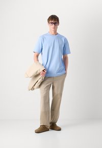 Ανοιχτό μπλε βαμβακερό μπλουζάκι με κοντά μανίκια, σε συνδυασμό με μπεζ παντελόνι που διαθέτει ραφές. Καφέ εσπαντρίγιες και ένα μπεζ μπουφάν κρεμασμένο σε έναν βραχίονά.