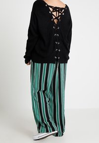 Pull en tricot noir avec un dos en V profond orné de liens croisés, associé à un pantalon large à rayures verticales vertes et noires.