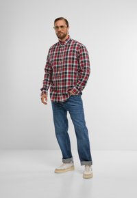 Karierte Hemd in Rot, Blau und Weiß; Button-down-Kragen, lange Ärmel. Kombiniert mit blauen Jeans und beigen Sneakern. Klare, lässige Gestaltung.